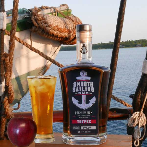 Smooth Sailing Rum bottle alongside a glass filled with Apple Juice, Smooth Sailing Rum on the rocks and an apple. In the background is one of the Tall Ship Picton Castle's dinghy with a fender made of weaved manila rope.